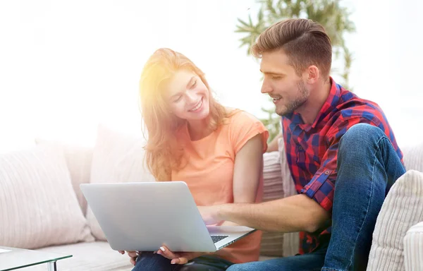 Joven pareja está utilizando un ordenador portátil y sonriendo mientras está sentado en el sofá —  Fotos de Stock