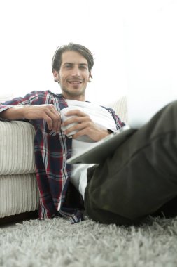 genç adam kanepede yakın katta oturan bir fincan tutan dizüstü bilgisayar ile