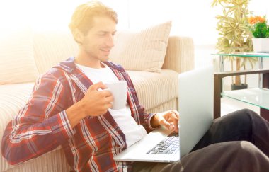 genç adam kanepede yakın katta oturan bir fincan tutan dizüstü bilgisayar ile