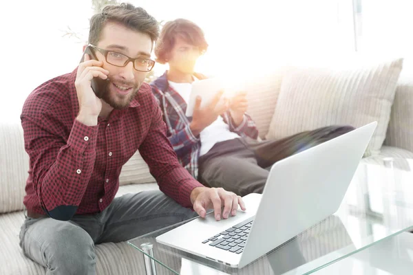 Man die op laptop werkt en praten over smartphone zittend op de Bank — Stockfoto