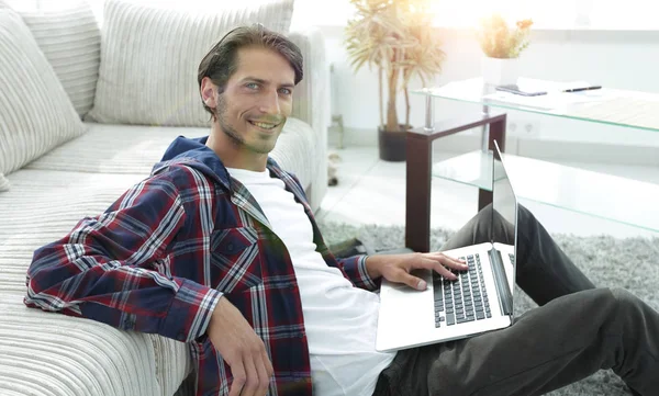 Jovem sorridente com laptop sentado no chão perto do sofá — Fotografia de Stock