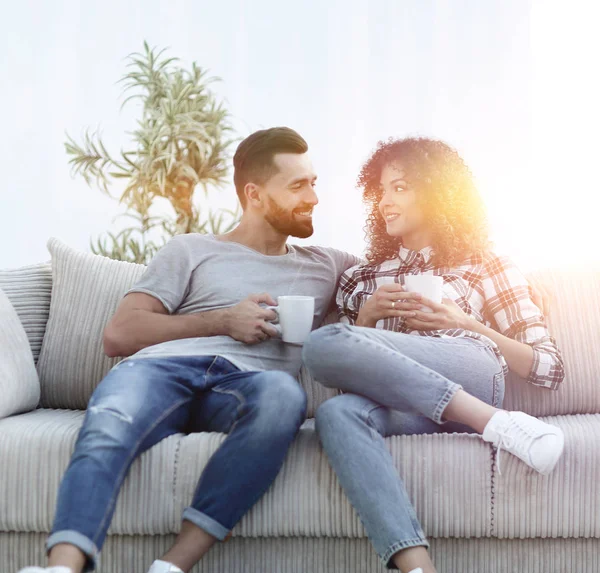 Hermosa pareja sosteniendo una taza de bebida mientras está sentada en el sofá en casa — Foto de Stock