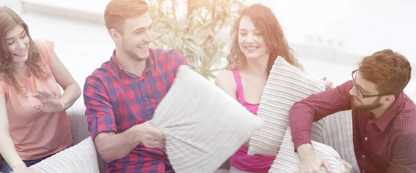 cheerful friends playing pillow fight, sitting on the couch