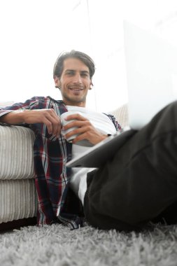 genç adam kanepede yakın katta oturan bir fincan tutan dizüstü bilgisayar ile