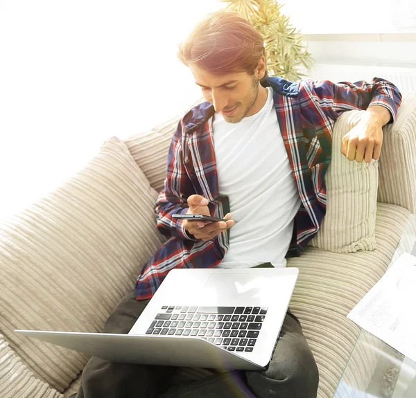Νεαρός άνδρας με laptop και smartphone που κάθεται στον καναπέ στο καθιστικό με συρόμενη — Φωτογραφία Αρχείου