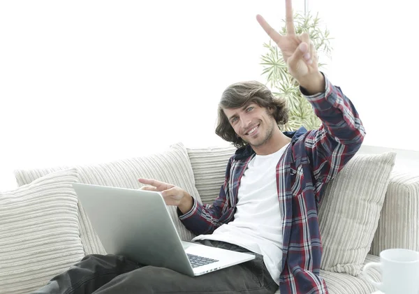 happy guy with laptop sitting on sofa and showing his hand a winning gesture