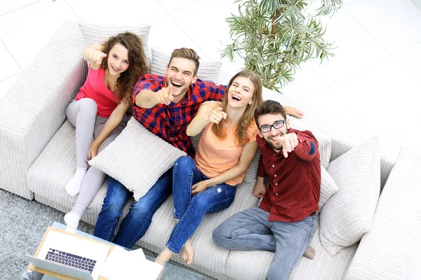 Grupo de amigos rindo sentado no sofá e mostrando indicador na câmera . — Fotografia de Stock