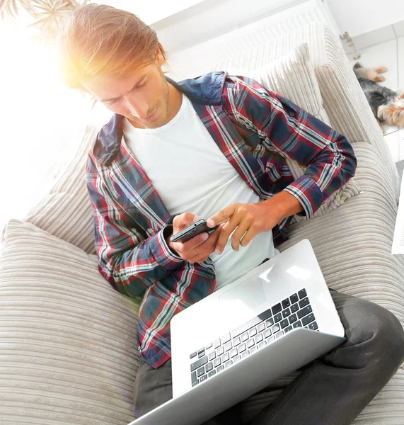 Jeune homme sérieux avec ordinateur portable et smartphone dans le salon . — Photo