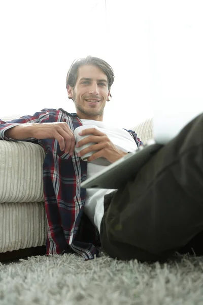 Joven con portátil sosteniendo una taza sentada en el suelo cerca del sofá —  Fotos de Stock