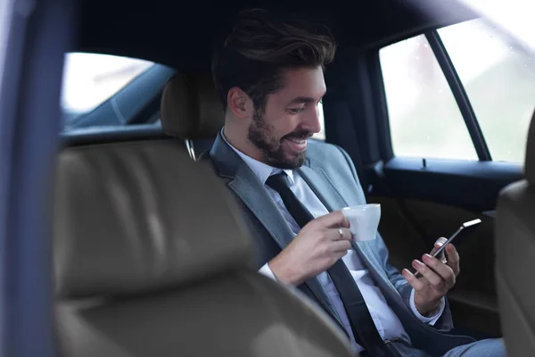 Бизнесмен пьет кофе в машине с телефоном в руке — стоковое фото