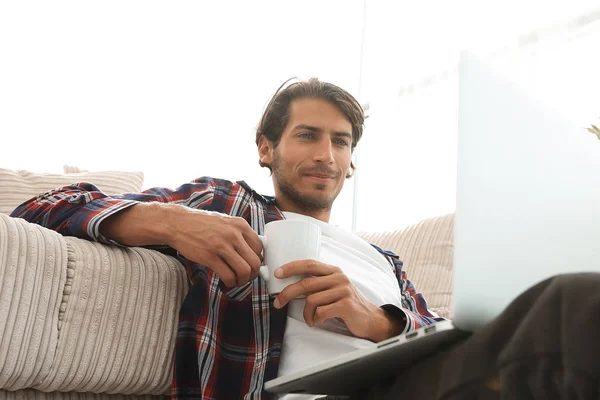 Jovem com laptop segurando uma xícara sentada no chão perto do sofá — Fotografia de Stock