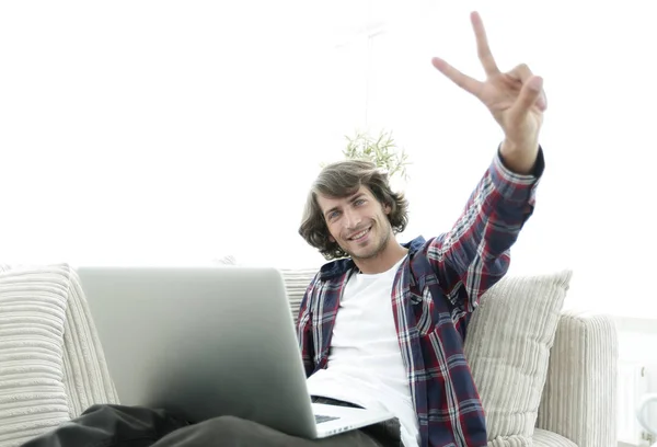 Gelukkig man met laptop zittend op een bank en toont zijn hand een winnende gebaar — Stockfoto