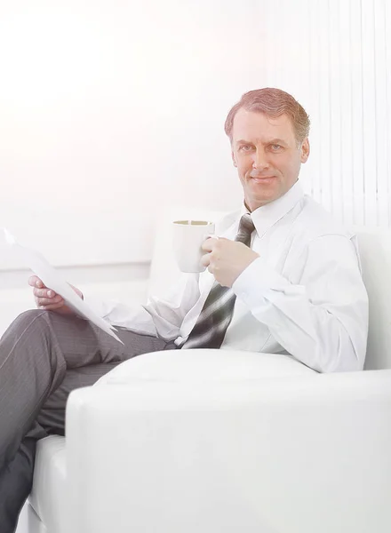 Confident businessman drinking tea and reading a document,sitting in a chair in the hotel room — Stock Photo, Image