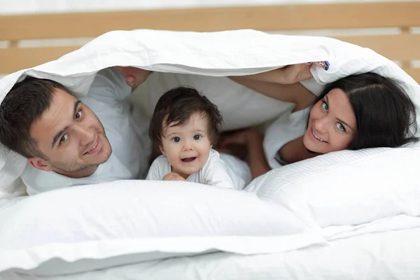 Familia feliz posando bajo un edredón mientras mira a la cámara —  Fotos de Stock