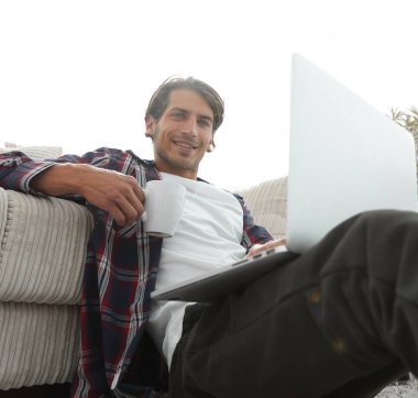 genç adam kanepede yakın katta oturan bir fincan tutan dizüstü bilgisayar ile