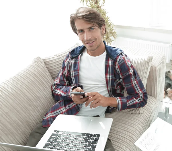 Joven serio con ordenador portátil y teléfono inteligente en la sala de estar . —  Fotos de Stock