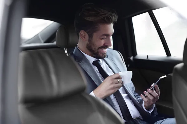 Uomo d'affari che beve caffè in auto con il telefono in mano — Foto Stock