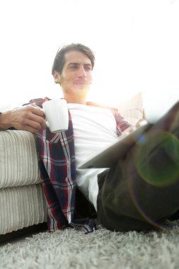 genç adam kanepede yakın katta oturan bir fincan tutan dizüstü bilgisayar ile