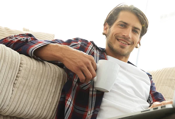 Ung man med laptop som håller en kopp som sitter på golvet nära soffan — Stockfoto