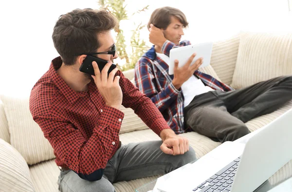 Cara trabalhando no laptop e falando no smartphone sentado no sofá — Fotografia de Stock