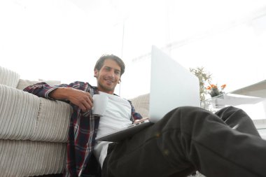 genç adam kanepede yakın katta oturan bir fincan tutan dizüstü bilgisayar ile