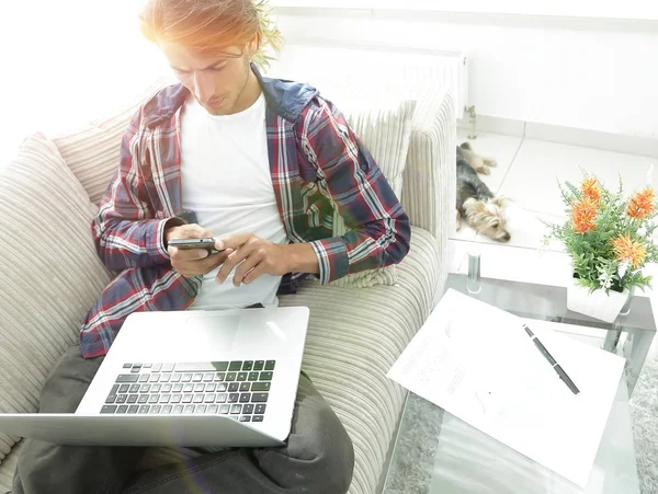 Joven serio con ordenador portátil y teléfono inteligente en la sala de estar . —  Fotos de Stock