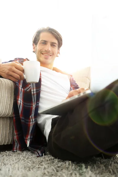 Jeune homme avec ordinateur portable tenant une tasse assise sur le sol près du canapé — Photo