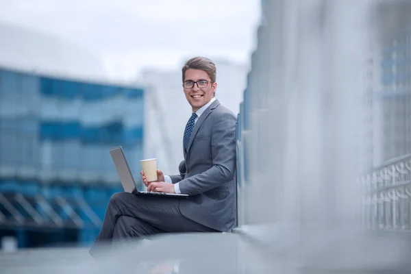 smiling corporate man with a take-away coffee, sitting next to an office building