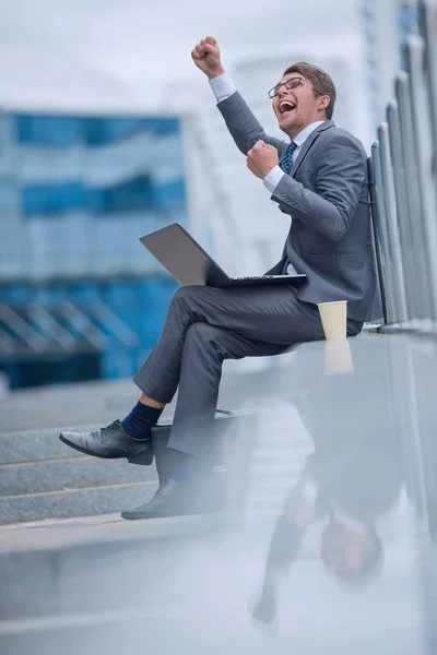 Hombre de negocios feliz con el ordenador portátil sentado cerca del edificio de oficinas . —  Fotos de Stock