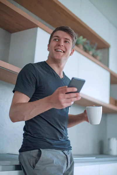 一个帅哥站在厨房里，手里拿着一杯咖啡和一部智能手机 — 图库照片