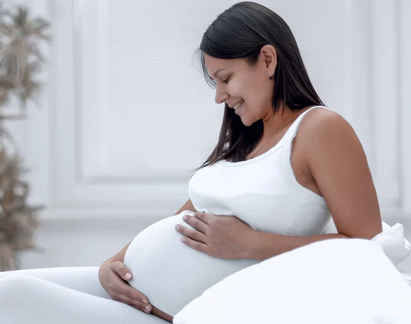 Vacker gravid kvinna sitter vid soffan och hålla handen på magen. — Stockfoto