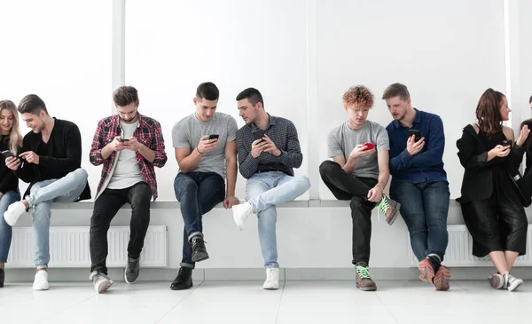 Grupo de jóvenes casuales con teléfonos inteligentes sentados en una fila . — Foto de Stock