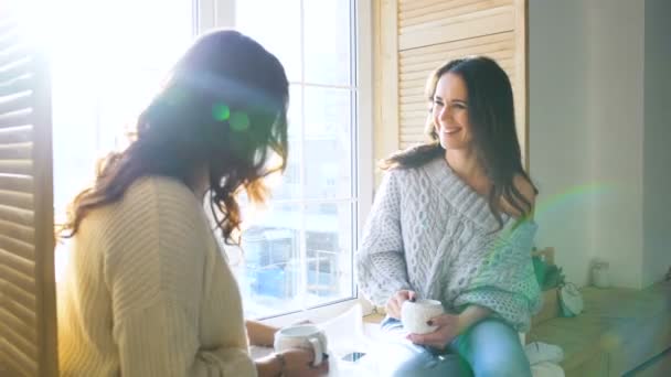Дві красиві дівчата обговорюють, сидячи на вікні. Подружки розважаються і сміються в спальні . — стокове відео