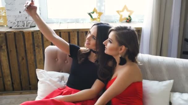 Two beautiful girls shoot selfie while sitting on sofa. Girlfriends having fun laugh in bedroom. — Stock Video