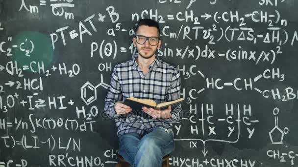 Joven científico hombre leer libro en ecuaciones químicas y matemáticas pared habitación interior — Vídeos de Stock