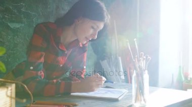 Genç kadın sanatçı resim gözleri üstünde kağıt defter kalem ile. Parlak güneş patlaması penceresinden