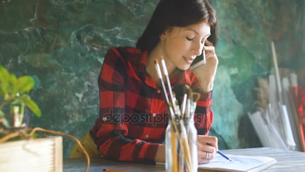 年轻女子艺术家绘画 scetch 在纸质笔记本和铅笔。女孩笑着谈电话 — 图库视频影像