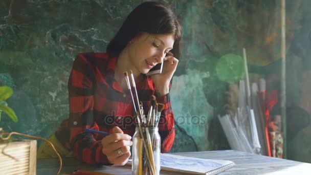 Joven artista pintando bocetos en cuaderno de papel con lápiz. Chica sonrisa y habla de teléfono — Vídeo de stock