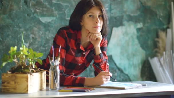 Young woman artist painting scetch on paper notebook with pencil and looks into camera smiling — Stock Video