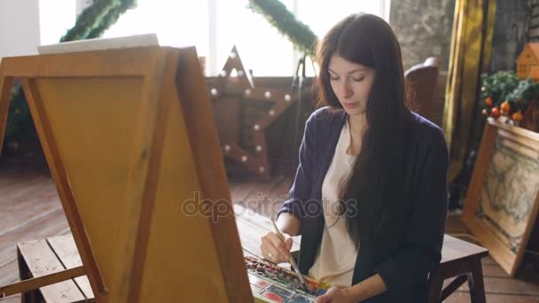 Νεαρή γυναίκα καλλιτέχνης επιστήσω ανέκδοτα με τα χρώματα watercolor και βούρτσα σε καβαλέτο καμβά — Αρχείο Βίντεο