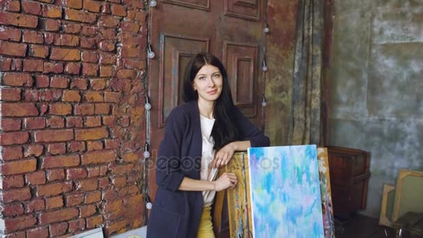 Молодая художница с акварелью стоит рядом с законченной картиной и улыбается в камеру — стоковое видео