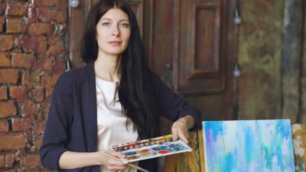 Молода жінка-художник з акварельними фарбами стоїть біля готової картини і посміхається в камеру — стокове відео