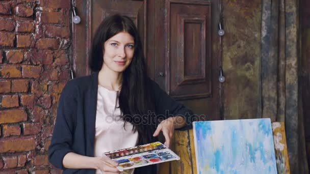 Медленный портрет молодой женщины-художника стоит рядом с законченной картиной и улыбается в камеру — стоковое видео