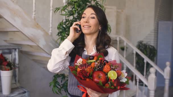 Kokkekvinneblomsterkiosk-telefon til klientene sine med blomster- og fruktblandet bukett – stockvideo