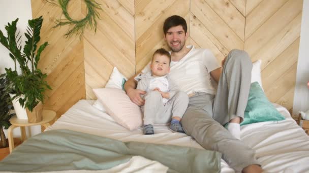 Retrato de feliz padre joven guapo y su hijo acostado en la cama sonriendo y posando en la cámara — Vídeo de stock