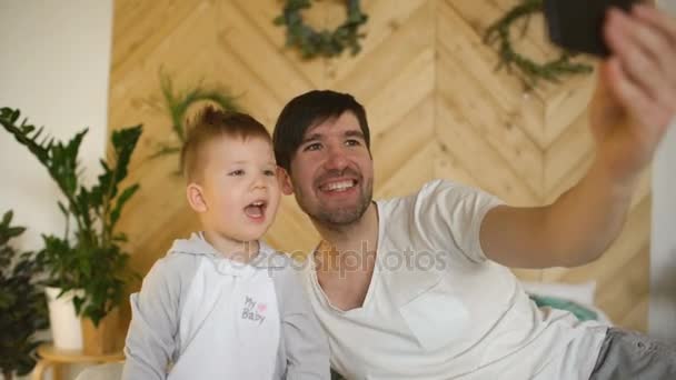 Young smiling father with son on bed taking selfie photo with smartphone camera — Stock Video