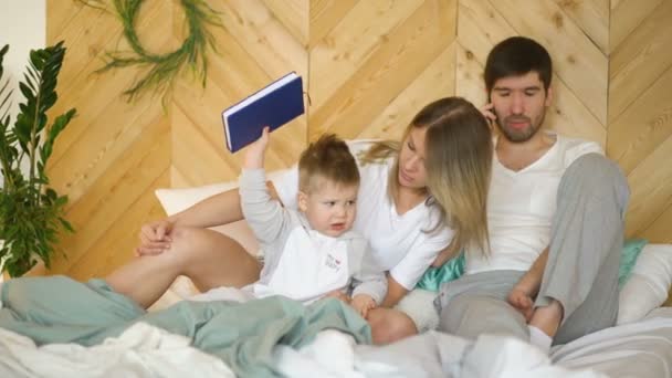 Young mother plays with son while father talking phone on bed — Stock Video