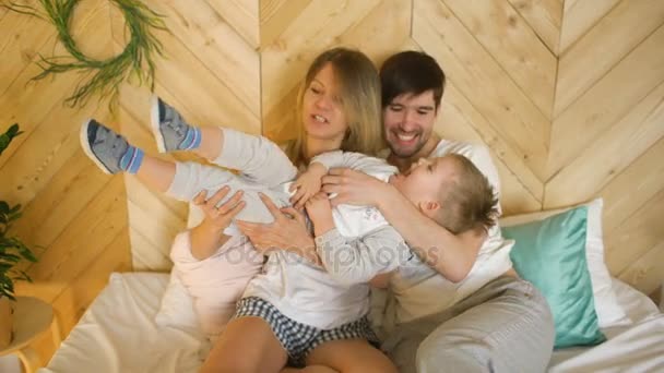 Una joven familia con hijo pequeño juega en la cama y lo besa sonriendo en el dormitorio — Vídeo de stock