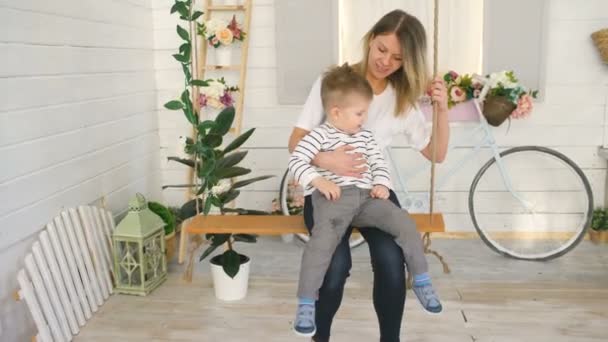 Gelukkig jonge moeder met haar zoontje zit op touw swing en spelen samen in hun slaapkamer — Stockvideo