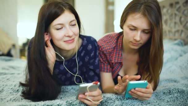 Dos amigas yuong escuchando música con auriculares conectados al teléfono inteligente yacen en la cama — Vídeo de stock